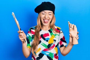 Kafkasyalı genç bir kadın sanatçı elinde küçük ahşap bir manikin ve boya fırçaları tutarken komik bir ifadeyle dilini dışarı çıkartıyor.. 