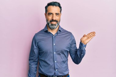 Orta yaşlı, sakallı, gri saçlı iş kıyafetleri giymiş neşeli bir şekilde gülümseyen ve avuç içi kameraya bakan bir adam.. 