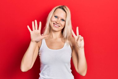 Güzel, beyaz tişört giyen beyaz tenli sarışın kadın, yedi numaralı parmaklarını gösterip gösterirken, kendinden emin ve mutlu bir şekilde gülümsüyor.. 