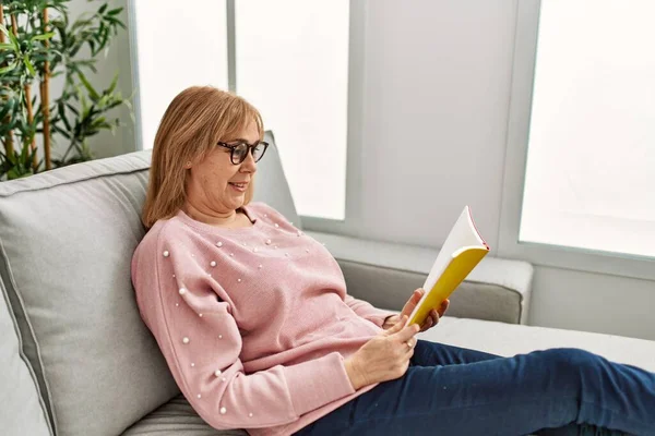 Medelålders Blond Kvinna Ler Glad Läsning Bok Liggande Soffan Hemma — Stockfoto