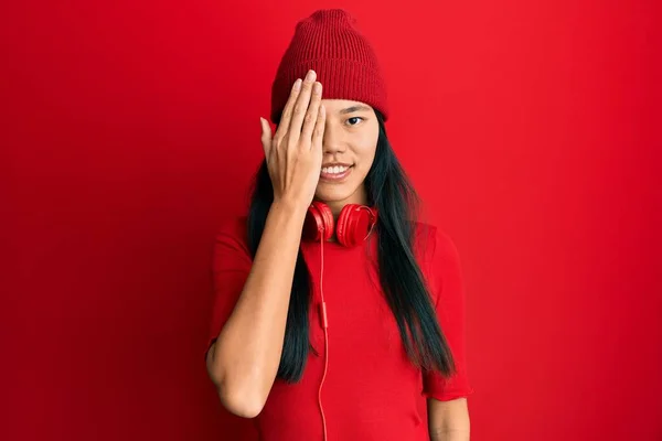 Young chinese woman listening to music using headphones covering one eye with hand, confident smile on face and surprise emotion.