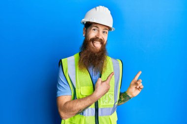 Uzun sakallı, güvenlik kaskı takan, yansıtıcı ceketli, gülümseyen ve kameraya bakan kızıl saçlı adam iki eliyle ve parmaklarıyla kenarı işaret ediyor.. 