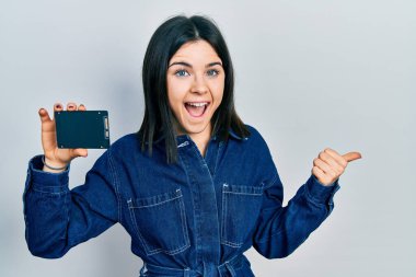 Genç esmer kadın SSD hafızasını tutuyor. Parmağını yana doğru gösteriyor. Ağzını açıp gülümsüyor. 