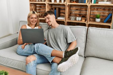 Evdeki kanepede oturup dizüstü bilgisayar kullanarak gülümseyen beyaz bir çift..