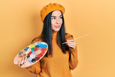 Boyalı fırça ve palet tutan genç esmer kadın. Bereli, gülümseyen, yan bakan ve düşünen.. 