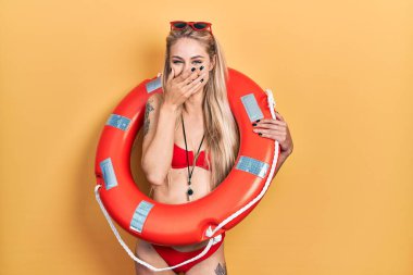 Bikini giyen, can kurtaran şamandırası tutan, gülen, utanan, ağzı ellerle kapatan, dedikodu ve skandal konsepti olan beyaz bir kadın. 