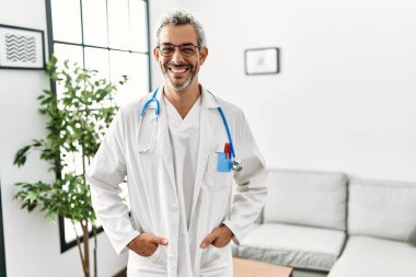 Orta yaşlı İspanyol adam doktor üniforması giyiyor ve bekleme odasında steteskop takıyor yüzünde mutlu ve havalı bir gülümsemeyle. şanslı kişi. 