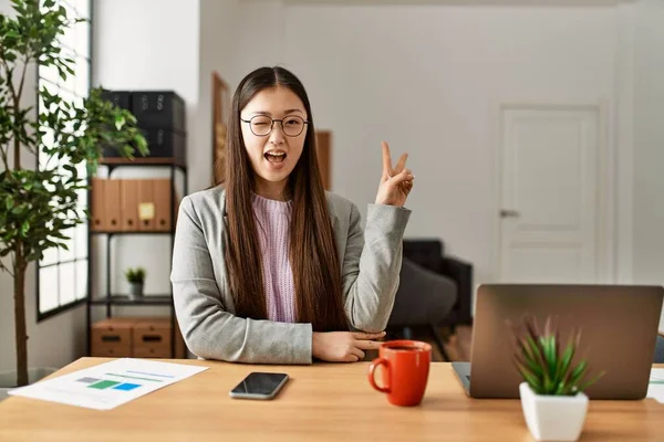 オフィスの机の上に座ってビジネススタイルを身に着けている若い中国のビジネスワーカーは 勝利のサインを行うカメラで幸せな顔をウィンクで笑っています — ストック写真