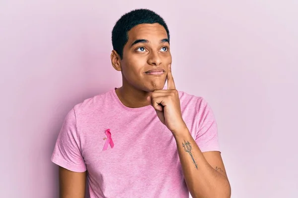 若いですハンサムなヒスパニック系の男身に着けていますピンク癌リボン上のシャツ深刻な顔考えます質問について手で顎 思慮深い混乱のアイデア — ストック写真
