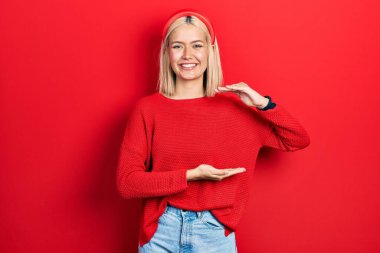 Güzel, sarışın bir kadın, sıradan kırmızı bir kazak giyiyor. Elleri büyük ve büyük boy işareti gösteriyor. Ölçüm sembolü. Kameraya bakıp gülümsüyor. ölçüm kavramı. 