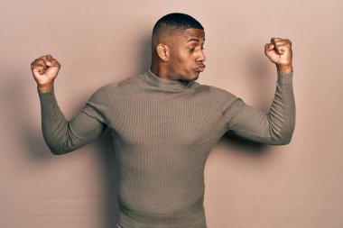 Balıkçı yaka kazak giyen genç bir zenci gururla gülümseyen kol kaslarını gösteriyor. fitness konsepti. 