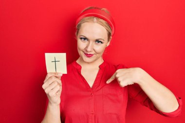 Elinde Katolik haçı tutan genç sarışın kadın mutlu ve gururlu bir kendini işaret ediyor. 