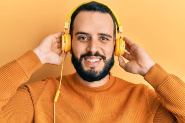 Sakallı genç adam kulaklık takarak müzik dinliyor mutlu ve havalı bir gülümsemeyle gülümsüyor. diş gösterme. 