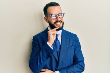 Sakallı, takım elbiseli ve kravatlı genç adam kameraya kendinden emin bakıyor kollarını kavuşturmuş ve çenesini kaldırmış bir şekilde gülümsüyor. Olumlu düşünmek. 