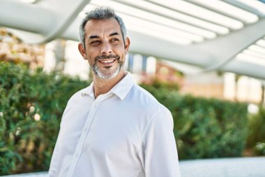 Orta yaşlı, kır saçlı, gülümseyen bir adam şehirde mutlu bir şekilde duruyor..