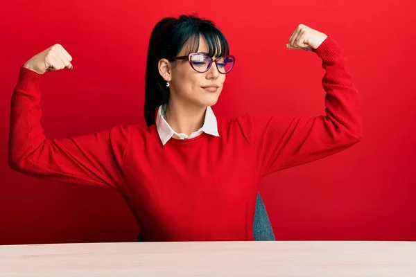 Young brunette woman with bangs wearing glasses sitting on the table showing arms muscles smiling proud. fitness concept.