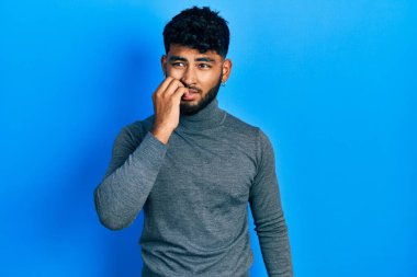 Balıkçı yaka kazak giyen sakallı bir Arap. Ağzı tırnak ısıran elleri yüzünden gergin ve stresli görünüyor. anksiyete. 