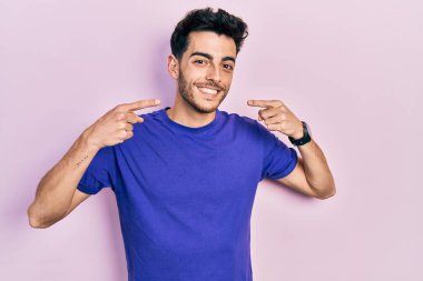 Gündelik tişört giyen, neşeli gülümseyen, parmaklarıyla ve ağzıyla işaret eden İspanyol genç bir adam. diş sağlığı kavramı. 