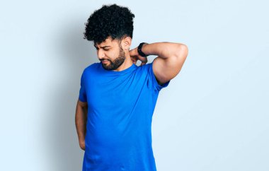 Sakallı genç bir Arap. Günlük mavi tişört giyiyor. Boyun ağrısı çekiyor, boynuna elle dokunuyor, kas ağrısı çekiyor. 