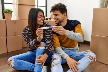 Genç Latin çift gülümsüyor. Mutlu hamilelik testi yapıyorlar. Yeni evlerinde yerde oturuyorlar..