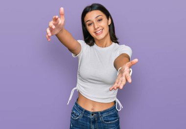 Latin asıllı genç bir kız, günlük beyaz tişört giyiyor kameraya bakıyor ve sarılmak için kollarını açmış gülümsüyor. Mutluluğu kucaklayan neşeli bir ifade.. 