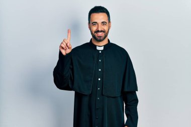 Sakallı, Katolik rahip cübbesi giyen yakışıklı İspanyol bir numarayı gösterip işaret ederken kendinden emin ve mutlu gülümsüyor.. 