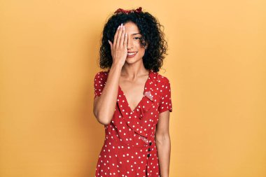 Yazlık elbise giyen genç Latin kız bir gözünü eliyle kapatıyor, yüzünde kendine güvenen bir gülümseme ve şaşırtıcı bir duygu var.. 