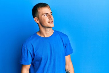 Günlük mavi tişört giyen, yüzünde gülümseme ve doğal ifadeyle yan yana bakan beyaz bir genç. Kendinden emin gülen. 