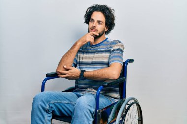Handsome hispanic man sitting on wheelchair with hand on chin thinking about question, pensive expression. smiling with thoughtful face. doubt concept.  clipart