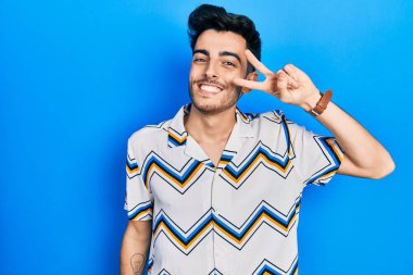 Gündelik kıyafetler giyen genç İspanyol adam barış sembolü yapıyor. Yüzünde parmaklar, gülümseyerek zaferi gösteriyor. 