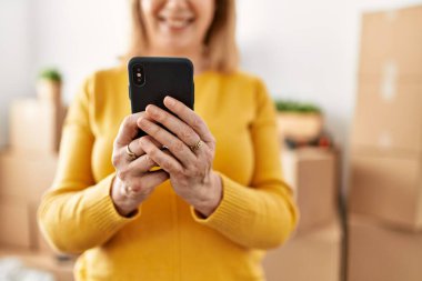 Orta yaşlı sarışın kadın yeni evinde akıllı telefon kullanarak mutlu bir şekilde gülümsüyor..