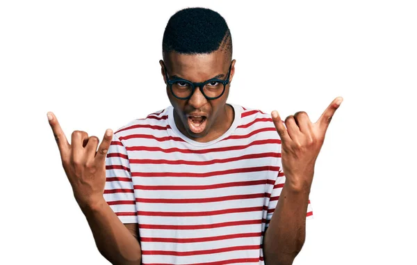 若いアフリカ系アメリカ人の男性は 手で岩のシンボルをやって狂気の表情で叫んでカジュアルなTシャツと眼鏡を着用しています 音楽スター 重い概念は — ストック写真