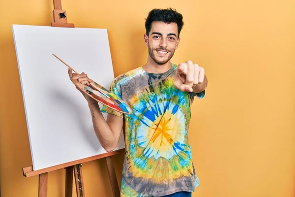 画家によってパレットで絵を描く若いヒスパニック系の男イーゼルスタンドはあなたと指でカメラを指しています — ストック写真