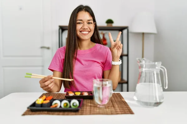 若いブルネットの女性が指で勝利サインをするカメラで顔がウィンクして幸せな笑顔で箸を使って寿司を食べています — ストック写真