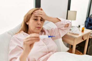 Orta yaşlı sarışın kadın. Sıcaklık ölçer. Evdeki yatağın üzerinde termometre tutuyor..