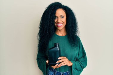 Orta yaşlı Afro-Amerikalı kadın elinde yağ şişesi tutarken pozitif ve mutlu görünüyor. Dişlerini gösteren bir gülümsemeyle ayakta duruyor. 