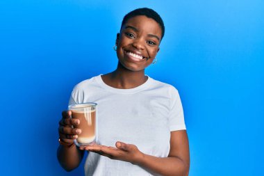 Afro-Amerikalı genç bir kadın elinde kahve tutarken pozitif ve mutlu görünüyor ve gülümsüyor. 
