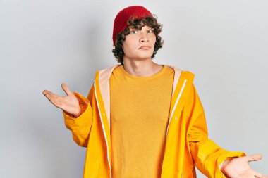 Sarı yağmurluk giyen yakışıklı ve elleri ve kolları havada, kafası karışmış genç bir adam. şüphe kavramı. 