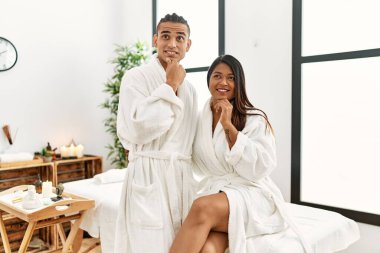 Havlu giymiş genç bir Latin çift güzellik merkezinde elleri çenelerinde soru sorarken düşünceli bir ifade takınıyorlar. Gülümseyen ve düşünceli bir yüz. şüphe kavramı. 