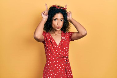Yazlık elbise giyen genç Latin kız komik jestler yapıyor. Parmağını boynuz gibi kafasının üstünde gezdiriyor. 