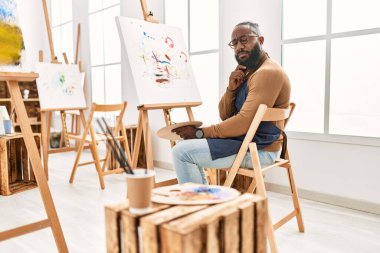 Afrikalı Amerikalı sanatçı, resim stüdyosunda tuvale resim yapıyor. Ciddi bir yüz ifadesi var. Elle çeneyi sorguluyor. 