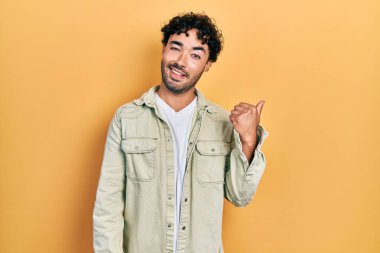 Gündelik kıyafetler giyen, gülümseyen, gülümseyen ve başparmağıyla yan tarafı gösteren İspanyol genç adam.. 