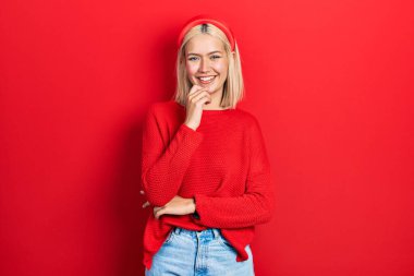 Güzel sarışın kadın, sıradan kırmızı bir kazak giyiyor kameraya bakıyor kollarını kavuşturup gülümseyerek ve çenesini kaldırarak. Olumlu düşünmek. 