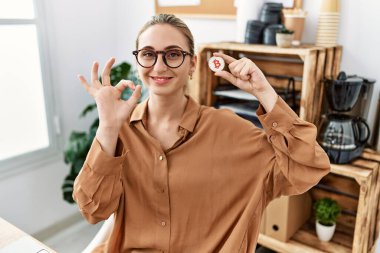 Ofiste elinde sanal para olan genç sarışın kadın parmaklarıyla imza atıyor, gülümseyerek mükemmel bir sembol oluşturuyor. 