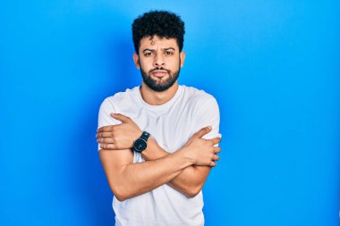 Sakallı, beyaz tişörtlü genç bir Arap kış soğuğu için titriyor ve donuyor. Yüzünde üzgün ve şok ifadesi var. 