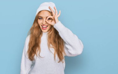 Genç beyaz kız yün kazak ve kış şapkası giyiyor. El hareketleriyle gülümsüyor ve mutlu yüzüyle parmaklarının arasından bakıyor.. 