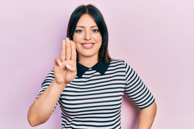 Genç İspanyol kadın gündelik kıyafetler giyer ve üç numaralı parmaklarıyla gülümseyerek mutlu ve kendinden emin bir şekilde işaret eder.. 