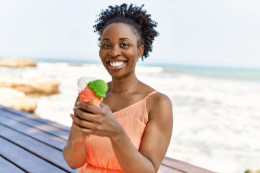Genç Afrikalı Amerikalı bir kadın kumsalda mutlu bir şekilde gülümsüyor ve dondurma yiyor.
