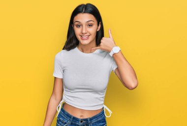 Latin asıllı genç bir kız, günlük beyaz tişörtler giyip gülümsüyor, telefonla el ve parmak hareketleri yapıyor, telefonda konuşuyor gibi. iletişim kavramları. 