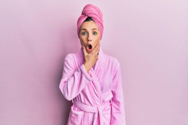 Duş başlığı ve bornoz takan genç sarışın kadın, elleri çenesinde, inanamama, şaşkınlık ve şaşkınlık dolu bir ifade takınıyor. 